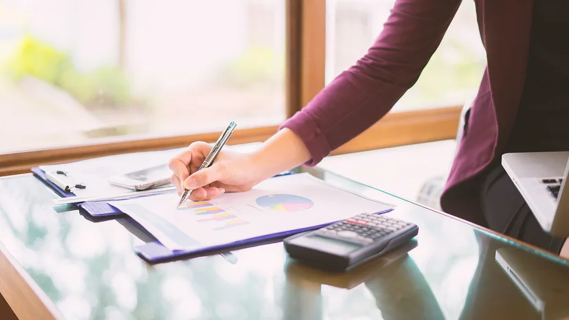 A person reviewing printed business charts with a pen beside a calculator and laptop