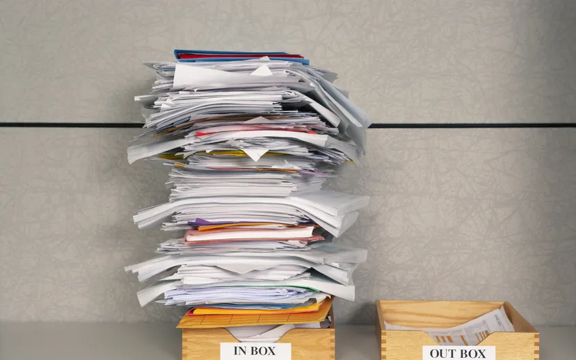 A tall stack of papers, folders and receipts piled on a wooden in-tray beside an almost empty out-tray.