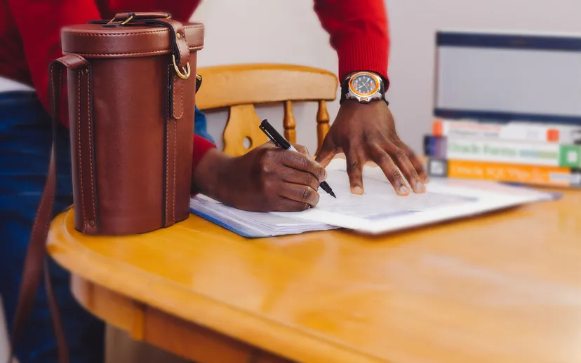 A person's hands filling in a printed form with a pen at a wooden table, face not shown.