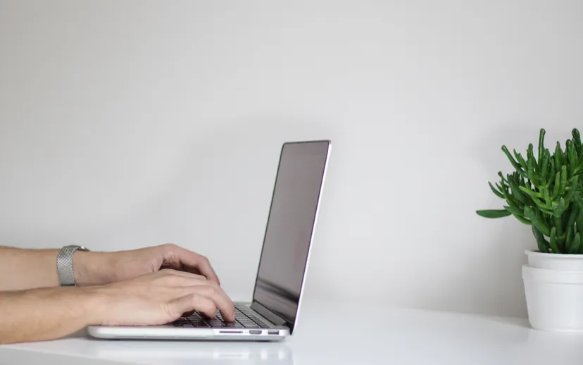 Hands typing on a laptop on a clean white desk next to a small potted plant.