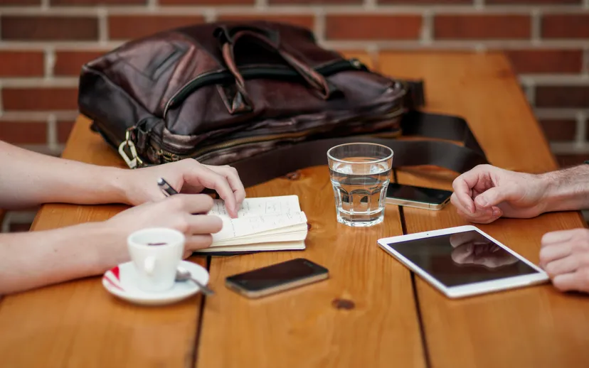 Two people meet at a wooden table with a notebook, coffee, phones and a tablet; only their hands are visible.