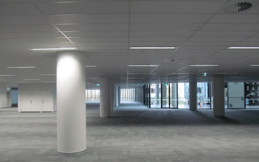 A large empty office floor with grey carpet, columns and windows, with no furniture.