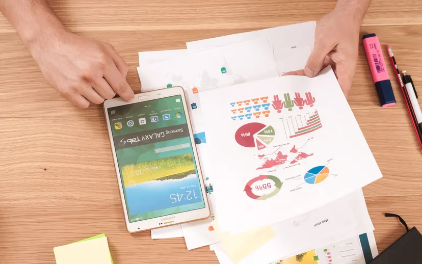 Hands arrange printed charts and reports next to a tablet, highlighter and pencils on a wooden desk.