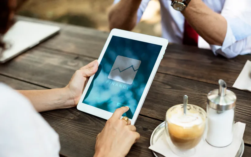 Hands holding a tablet showing a finance chart beside a cup of coffee on a wooden table.
