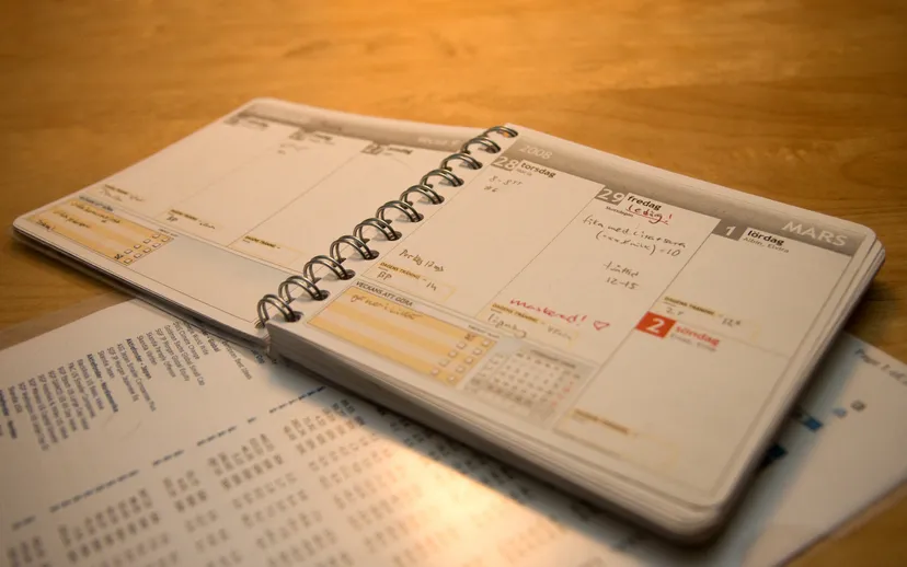 A spiral-bound weekly planner with handwritten notes lies open on a wooden desk.