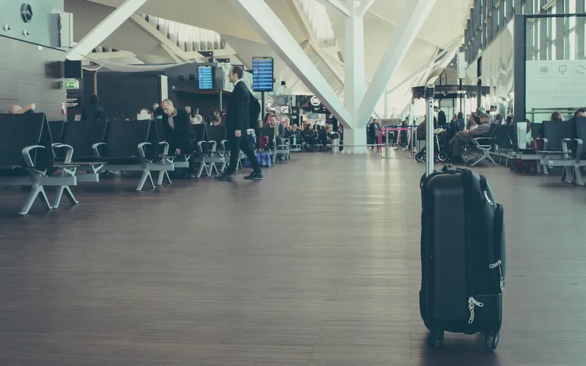 A black suitcase standing in a bright airport departure lounge with travellers in the background.