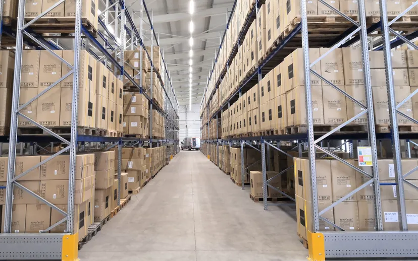 A long aisle runs between tall racks of boxed goods in a bright warehouse.