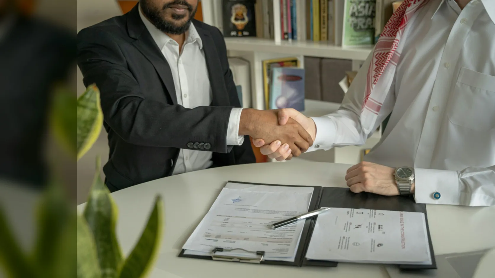 Two businessmen, one in Gulf dress, shaking hands over signed documents